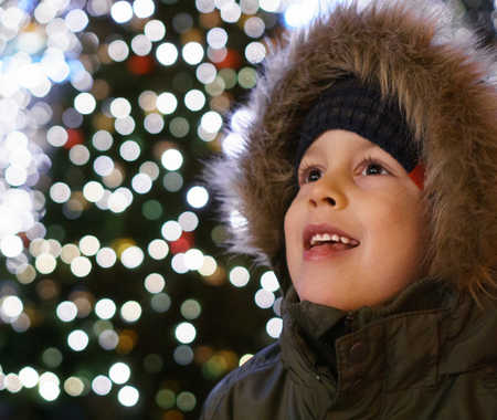A child in a fur hat smiles as they stand in front of a Christmas tree with white lights.